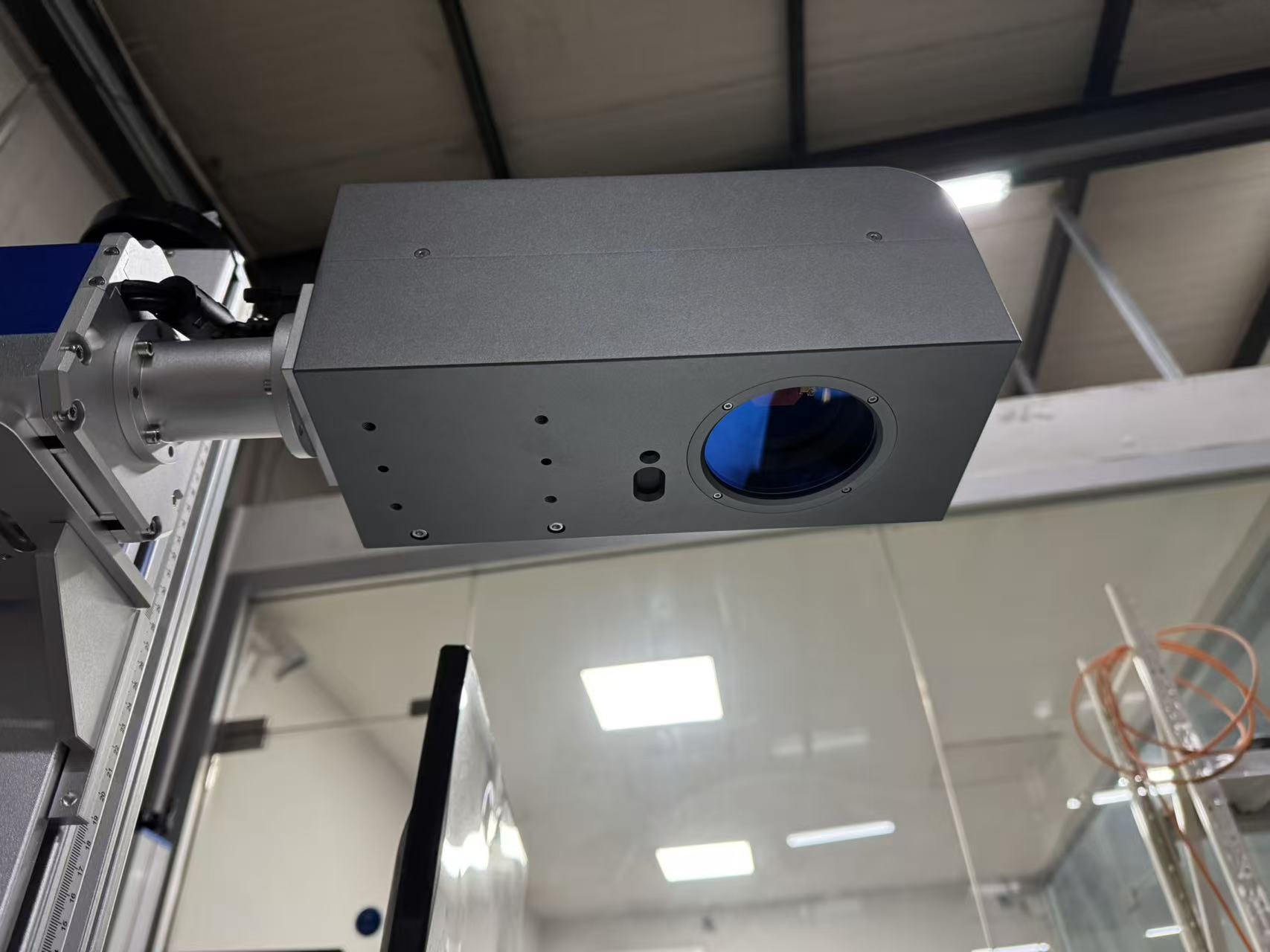 Gray rectangular industrial enclosure with a circular blue-tinted viewport, mounted on a rail/shaft in a factory setting.