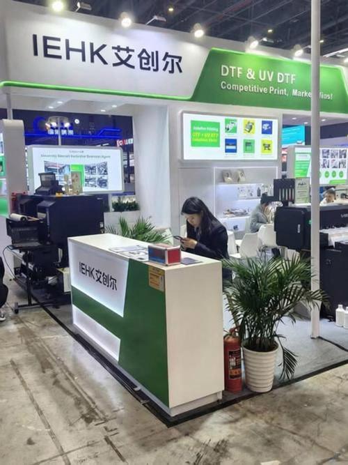 Exhibition booth for IEHK with green and white signage, a receptionist desk, and plants at a trade show. Two staff work at computers in the background.