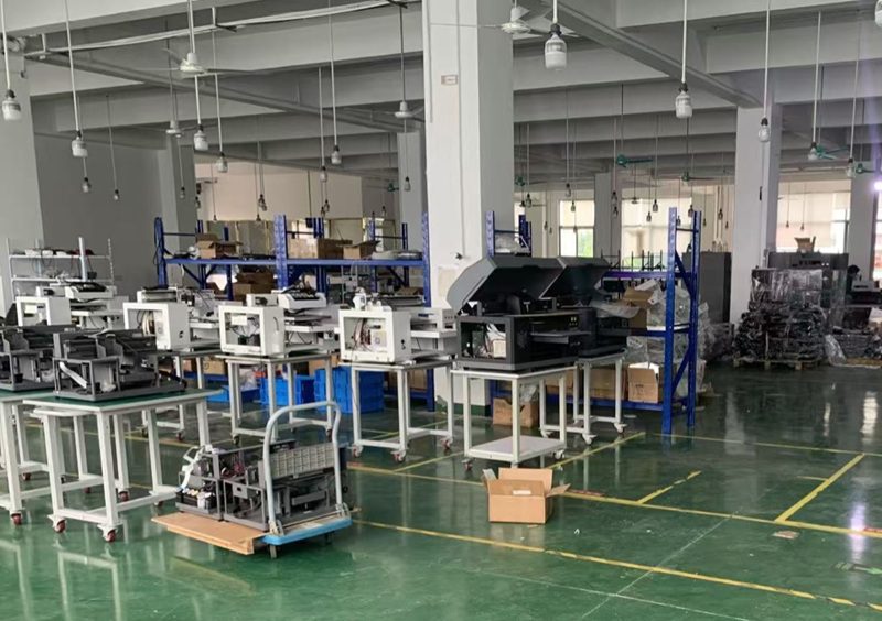 Factory floor with multiple CNC/printing machines on metal benches and blue storage racks, cardboard boxes nearby.